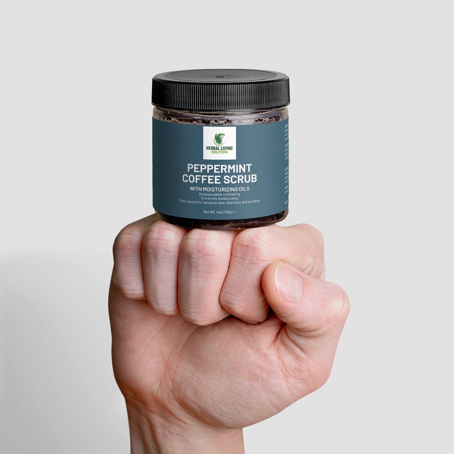 Hand holding a jar of Peppermint Coffee Scrub with moisturizing oils against a gray background